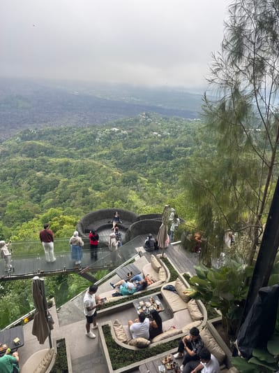 The image depicts an outdoor seating area of a restaurant or cafe perched on a cliffside, offering a panoramic view of a lush, green valley and a misty, volcanic landscape in the distance. In the foreground, several groups of people are visible. On a raised wooden deck, a group of individuals are seated on large, curved beige sofas, some with drinks on low tables. A man in a white t-shirt and black shorts stands in the center of this deck, looking at his phone. Further back, on a transparent walkway extending over the cliff edge, a few people are standing, observing the view. One woman in a black dress is seated on a large, woven circular sofa, also overlooking the valley. The overall environment suggests a scenic tourist spot in Kintamani, Indonesia, known for its volcanic caldera views. The weather appears to be overcast, contributing to the misty atmosphere.