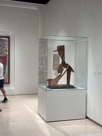 The image depicts an interior scene within the Picasso Museum Málaga, located in the city of Málaga, Spain. The focal point is a wooden sculpture displayed inside a glass vitrine. The sculpture, abstract and stylized, resembles a bird-like figure with elongated features and a prominent beak-like protrusion. It rests on a dark base within the protective display case, which is illuminated by soft overhead lighting.

To the left of the sculpture, a framed painting is visible, featuring abstract shapes and lines in various colors. A person, seen from the waist down, is walking past the painting on the left side of the frame. This individual is wearing dark shorts and a white t-shirt, and dark sneakers. The floor is tiled with a pale, polished stone, reflecting the light. The walls are a clean, neutral white, creating a minimalist gallery ambiance. Two small informational plaques are affixed to the wall to the right of the sculpture's display case. The overall environment suggests a museum setting, focused on the exhibition of art.