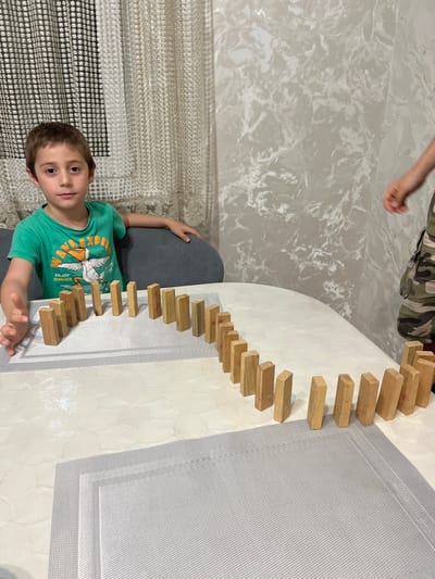 The image depicts a young boy, likely between 5 and 7 years old, sitting at a table. He is wearing a green t-shirt with a graphic print of a pelican and the words "ENJOY SUMMER." His left arm is extended and slightly blurred, suggesting he might be in motion or about to interact with the dominoes. The table is covered with a white tablecloth and placemats. A line of wooden dominoes is set up on the table, forming a curving path. In the background, a second person, partially visible and wearing camouflage shorts, has their right arm extended towards the dominoes, implying they may also be involved in the activity. The room is furnished with curtains and patterned wallpaper. The context of Kaspiysk, Russia, is not ascertainable from the visual information provided in the image.