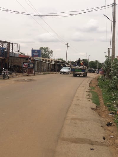 The image shows a street scene in Hanwa, Nigeria. A truck carrying people and goods is traveling on the road. A white car follows behind. The road is paved. On the left side of the road are buildings, including shops, and signs for "Dangote Cement" and "Abtech POS". A motorcycle is parked near one of the buildings. Overhead power lines run above the street. The sky is overcast. Vegetation grows along the roadside on the right. The scene suggests a bustling commercial area.