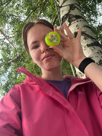 A person, possibly a woman, is shown from a low-angle perspective, looking up towards the camera. They are wearing a bright pink hooded jacket over a purple shirt. A black wristband is visible on their left wrist. In their right hand, they hold a small, round, green container with a label. The person's face is partially obscured by the container.
The background consists of a large birch tree with white bark and dark markings. Lush green leaves and branches from the tree, as well as other trees, fill the upper portion of the frame, suggesting an outdoor setting in a wooded area. The lighting appears to be natural daylight, likely daytime, with dappled light filtering through the leaves. The overall mood seems relaxed and casual.
The label on the green container is partially visible and appears to be in Cyrillic script. Text that can be identified includes "СПЛАТ" (SPLAT) and "ЗУБНАЯ НИТЬ" (dental floss). Other text is too small to be legible.
The location context provided is Sergino, Russia. This suggests the image was taken in a natural environment within that region. The image appears to be a selfie or a candid shot taken outdoors.