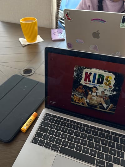 Two open Apple laptops sit on a table. The first laptop is silver and covered in stickers, featuring an Apple logo. The second laptop shows the album "K.I.D.S." by Mac Miller on the screen, with the keyboard visible. A yellow cup rests on a napkin to the side. A purple object and a dark gray tablet case are also present. A tube of lip balm sits near the keyboard.