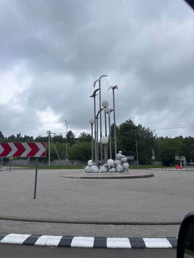 The image depicts a roundabout in the city of Zhdanovichsky Selsoviet, Belarus. The roundabout features a circular sculpture in its center, composed of several tall metal poles with stylized bird figures perched on top. Spherical elements are clustered at the base of the poles. The roundabout itself is paved with interlocking grey bricks. Traffic signs, including a directional sign with red and white chevron stripes, and a pedestrian crossing sign with a yellow triangle and a black pedestrian silhouette, are visible. The background consists of trees and a cloudy sky.