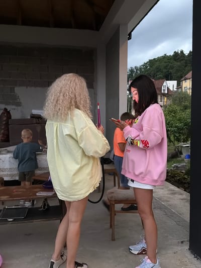 The image depicts an outdoor scene, likely a covered porch or patio, with a rural or suburban backdrop suggesting the outskirts of Baranovka, Russia. The environment is characterized by a partially constructed brick wall on the left and a concrete floor. In the foreground, two young women are the primary subjects. The woman on the left has voluminous blonde curly hair and is wearing a loose, pale yellow shirt. She is holding a pink object, possibly a vape pen or a small tool. The woman on the right, with dark, straight hair, is wearing a pink oversized sweatshirt with colorful graphic details on the sleeves and a white skirt. She is engrossed in her mobile phone, holding it in her hands and looking down at the screen.

In the mid-ground, a young boy wearing a blue t-shirt and dark shorts is standing and appears to be interacting with something on a table covered with a white tablecloth. Another individual, wearing an orange t-shirt, is partially visible behind the women. The background reveals a residential area with houses and lush green trees under a cloudy sky. A wooden table and chairs are also present, indicating a casual gathering or social setting. The overall impression is one of everyday life in a semi-urban or rural Russian locality.