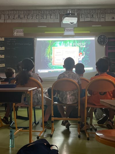 The image depicts a classroom setting in La Fouillouse, France, where a group of young students are gathered around a projector screen. The students are seated on yellow chairs, facing the screen, and appear to be engaged in a lesson. The projector displays an image with the words "PIANO FOREST" prominently featured, along with text in Japanese and French, suggesting a lesson related to language or music.
Several objects are visible in the classroom, including a wall clock on the upper left, a small white projector or scanner device, and a blackboard with handwritten notes. A numbering system from 19 to 63 is displayed vertically on the wall to the left of the projector screen. The overall lighting suggests it is daytime.
The students' posture and focus on the screen indicate they are attentive. One student in the foreground is looking down at a notebook. The atmosphere appears calm and focused, typical of a classroom environment. The chairs are simple, tubular metal frames with yellow seats and backs. The walls are painted a light green. The scene captures a moment of learning and engagement in a French educational setting.