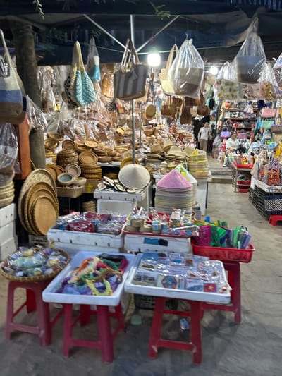 The image depicts a busy marketplace stall, likely in Hội An Ward, Vietnam, characterized by an abundance of handcrafted goods. In the foreground, a red plastic stool supports a woven tray filled with various colorful beads and necklaces. Adjacent to it, another red plastic stool holds a white rectangular tray overflowing with similar colorful beaded jewelry. Further to the right, a third red stool displays a white rectangular tray covered in plastic, revealing a collection of small, packaged items, possibly souvenirs.

Behind these foreground displays, more stalls are visible, laden with an assortment of items. Stacks of woven hats and baskets are prominent, as are numerous bags of various designs and materials, some hanging from the ceiling and others displayed on shelves. The overall impression is of a vibrant and crowded market, with goods neatly arranged for sale. In the mid-ground, two individuals are partially visible, one appearing to be a vendor in a white shirt and the other a woman in a white shirt. The lighting suggests it might be evening, with artificial lights illuminating the scene. The ground appears to be concrete.