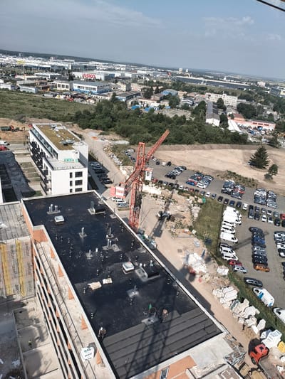 This is an aerial drone shot of a construction site in Pilsen, Czechia, with an industrial area in the background. The foreground shows a multi-story building under construction with workers on its roof. There are also several other buildings in various stages of construction, along with construction materials, equipment, and vehicles. In the background, there is a large industrial park with many warehouses and commercial buildings. The sky is clear and blue with a few scattered clouds.