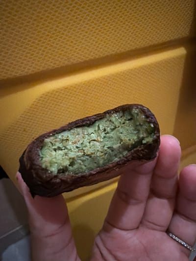 The image shows a person holding a partially eaten food item. The food item appears to be a dark brown outer shell with a light green, textured filling. It resembles a type of cookie, pastry, or energy bar that has been bitten into. The person's hand, which is holding the food, has a ring with small, clear stones on one finger. The background is a textured yellow surface, possibly a placemat or tray, with a shadow casting a diagonal line across it. The lighting suggests an indoor setting. The image is a close-up shot, focusing on the food item and the hand. The scene is simple and likely depicts a casual moment of someone eating or examining their food. There is no visible text in the image. The color palette is dominated by brown, green, and yellow.