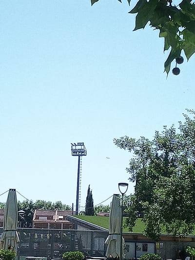 The image depicts an outdoor scene under a clear blue sky. In the foreground, two large, light-colored umbrellas are partially visible, one on the left and another on the right. Behind these umbrellas, a building with a red-tiled roof is visible. Signs with letters, potentially spelling out "BANYOLES," are mounted on a railing in front of this building.

Further in the background, a tall, slender pole with a structure at its top, likely a lighting tower or a communications mast, stands prominently against the sky. A thin, dark tree with a conical shape is situated to the right of this tower. A large, dense tree with lush green foliage dominates the right side of the image, its branches extending towards the sky and partially obscuring the view. A street lamp with a distinctive curved shade is visible near the base of this tree.

The overall environment appears to be an urban or semi-urban setting, possibly a park, plaza, or outdoor dining area, given the presence of umbrellas and seating structures suggested by the blurred elements in the lower part of the image. The presence of the sign with "BANYOLES" suggests the location is the city of Banyoles, Spain.