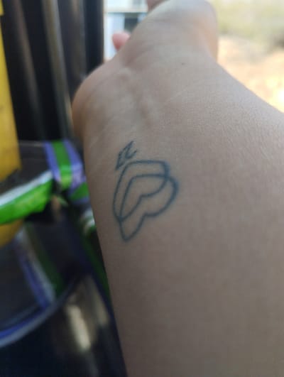 The image shows a close-up of a person's wrist with a simple tattoo. The tattoo is a line drawing of what appears to be two overlapping heart shapes with a small diamond-like shape above them. The skin is light-toned. The wrist is partially visible, with the rest of the arm extending out of frame. The background is blurred, but includes a glimpse of green and blue stripes, and possibly other objects. The location, Punta de Piedras, Venezuela is not evident from the visual content. No people, actions, or interactions are discernible beyond the presence of the tattoo on the wrist. The environment visible is limited and indistinct.