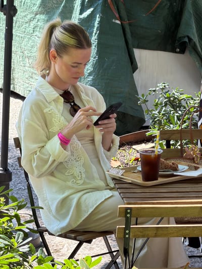 Captured from an overhead perspective, the image portrays a woman seated at a slatted wooden table in what appears to be an outdoor setting, possibly in Bunyola, Spain. The woman is fair-skinned with blonde hair styled in a low ponytail, and she's engaged with her smartphone, held in her left hand. She is dressed in light-colored clothing, including a white shirt with lace detailing and a matching dress. A pink wristband adorns her left wrist, with the words "TOILET CODE" and a number "123456#" printed on it. A pair of sunglasses rests on her shirt, close to her collar.
The table in front of her holds a tray with a beverage in a clear plastic cup, complete with a straw, and what seems to be a pastry. Greenery is also part of the table setting, and foliage is visible on the periphery of the image. A green tarp is seen in the backdrop, providing a shaded area. The seating consists of a metal-framed chair that appears to be part of an outdoor dining set. The ground beneath the chair seems to be paved or made of stone.