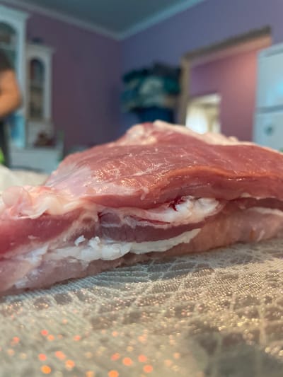 The image shows a close-up of a piece of raw pork, likely pork belly, resting on a crocheted lace cloth. The pork has layers of fat and lean meat. There is a small amount of blood visible within the fat layers. The background includes a blurred view of what appears to be a room interior with a purple wall and a shelf. The image does not contain any information about location in Olginka, Russia or any people, subjects, or actions taking place.