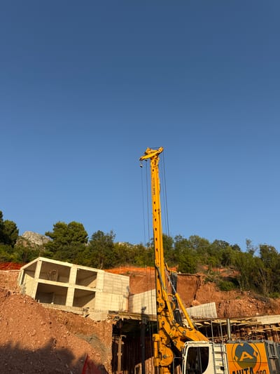 This media file depicts a construction site under a clear blue sky, likely during daylight hours given the bright illumination. The scene is dominated by a large yellow drilling rig, identified by the text "BAUER BG 23 H" on its boom. The rig is positioned amidst a hilly, wooded terrain, suggesting an outdoor, possibly mountainous or coastal, environment.

In the foreground and mid-ground, the construction is evident with exposed concrete structures and piles of reddish-brown soil. A partially built concrete building with visible internal framing is a prominent feature. Construction fencing, some of it orange netting, encloses parts of the site. The backdrop includes dense green trees and rocky hills, characteristic of a Mediterranean landscape. The text "ANITA 200" is visible on the side of the drilling rig's cabin, likely its model or company designation. The overall impression is one of ongoing infrastructure development in a natural setting.