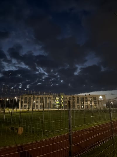 The image captures a wide shot of a building at dusk in Naberezhnye Chelny, Russia, under a cloudy sky. In the foreground, a chain-link fence encloses a grassy area, which appears to be a sports field or park. Beyond the fence, a multi-story building with numerous windows is visible, illuminated from within. The building's facade is constructed of brick-like material. A logo, a white triangle within a circle, is prominently displayed on the building's upper section, accompanied by text that appears to be in Cyrillic. The overall atmosphere is subdued due to the low light conditions.