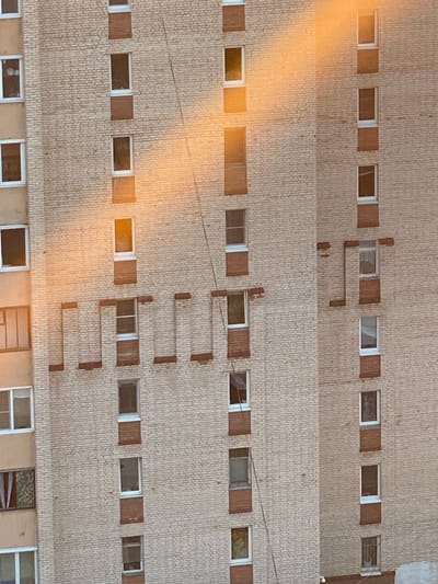 The image shows a section of a multi-story brick building. The building has many windows, some of which are open. The brickwork is light-colored with darker mortar. A bright, orange-yellow light streaks diagonally across the upper portion of the building, suggesting sunlight or a similar bright light source. The scene is likely an exterior view of an apartment building. There are no people or animals visible in the image. The environment appears to be urban.