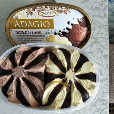In Cupcini, Moldova, an overhead shot captures a large, oval-shaped tub of Sandra brand "ADAGIO" Chocolate & Banana ice cream. The lid, partially visible, features golden text and imagery of cocoa pods and bananas, along with images of chocolate ice cream and sauce. Below the lid, the ice cream itself is presented in a clear plastic container, revealing a swirled pattern of chocolate and vanilla ice cream, interjected with dark chocolate sauce. The ice cream has a swirled, almost floral design radiating from the center. The container sits on a marble-patterned surface.
