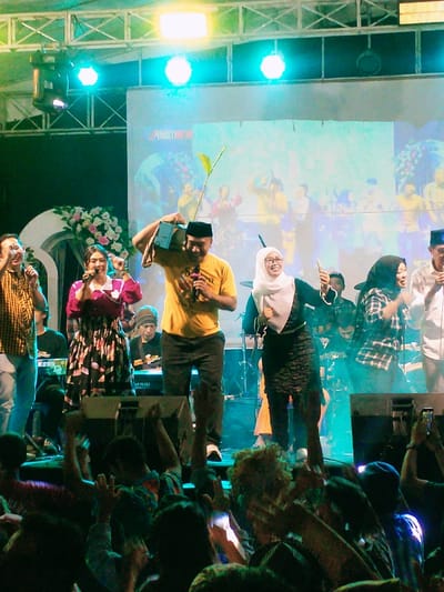 The image shows a stage with several performers in front of a large audience. The stage is illuminated by bright blue spotlights.  A man in a yellow shirt is holding a television set above his head. A woman in a black dress and white headscarf is holding a microphone in her right hand and dancing. Behind the performers is a large screen with the word "MULTI" visible.  The audience is cheering and raising their hands. The scene is set in the city of Kecamatan Sambirejo, Indonesia (latitude: -7.501, longitude: 111.088).