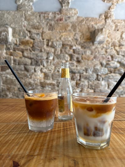 This is a close-up, eye-level shot of two iced drinks and a bottle of tonic water on a wooden table. The drinks are in clear glasses, one on the left with a dark liquid topped with ice and a slice of orange, and another on the right with layers of white and brown liquid, also with ice. Both drinks have black straws. A clear glass bottle with a yellow and white label is positioned between the two drinks, with some tonic water remaining inside. The background is a rough, textured stone wall, likely in Pisa, Italy, with the texture of the individual stones clearly visible. The lighting is soft, creating a warm ambiance. No people are visible in the image.