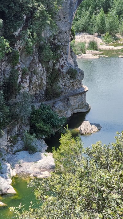 The image depicts a scenic view of the Pont del Liers, an ancient stone bridge spanning the river Muga in Tortellà, Spain. The bridge's weathered stone arches and masonry are visible, partially obscured by lush green vegetation and trees that cling to the rocky cliff face. A narrow pathway or road appears to follow the curve of the bridge, hugging the rock. The clear, calm waters of the river Muga are seen below, reflecting the surrounding landscape and the bright sky. The scene is bathed in natural sunlight, creating a serene and picturesque atmosphere.