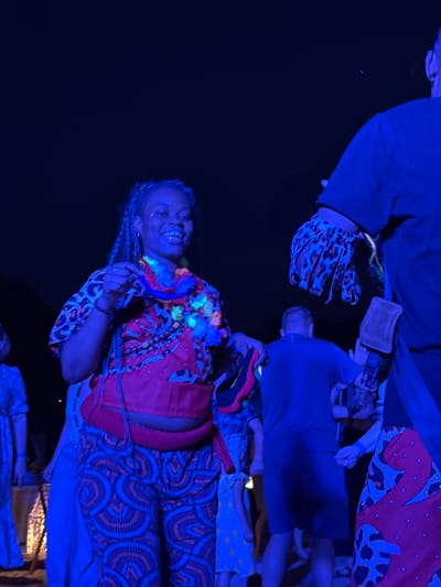 The image shows a group of people in what appears to be an outdoor evening event, possibly a festival or party, illuminated by blue and pink lights. In the foreground, a Black woman with braided hair is smiling and looking towards the camera. She is wearing a red top with a black and brown pattern, a colorful lei around her neck, and patterned pants. To her right, the arm of another person is visible, wearing a glove with a leopard print pattern. In the background, several other individuals are present, some dancing or moving, illuminated by the colored lights. The scene suggests a lively and festive atmosphere. The specific location in "городское поселение Щелкино, Ukraine" cannot be determined from the visual information alone.