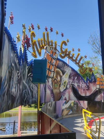 This is a photograph of a carnival ride on a bright, sunny day. The ride features a vibrant mural with graffiti-style artwork depicting what appears to be a stylized face with green lips and an eye. The ride itself is decorated with numerous strings of colorful lights in red, yellow, and blue, arranged in star and spherical shapes. A large, blue rectangular box is attached to the ride, possibly a speaker or control unit, connected by a yellow pole.

The background shows a clear blue sky and some green trees, suggesting an outdoor amusement park or fairground. In the distance, a body of water, likely a lake or pond, is visible, bordered by a decorative fence. The overall impression is one of excitement and festivity, typical of a carnival atmosphere. The time of day is clearly daytime, given the bright sunlight and distinct shadows cast by the ride's structure. The location cues, like the fence and the water, are consistent with a park or recreational area. No text is clearly legible on the ride or its surroundings.