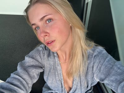 The image presents a close-up portrait of a single female individual, observed within an indeterminate indoor or sheltered outdoor setting in Skanderborg, Denmark. The subject, a young adult with fair skin, blue eyes, and long blonde hair, directly faces the camera. Her face shows a slight natural flush on the cheeks, and her lips are slightly parted. She wears a light blue-grey, textured, long-sleeved shirt, unbuttoned to partially expose a dark undergarment. Accessories include a thin gold-colored necklace on her collarbone and a small, square earring in her visible right ear. The background features a dark grey textured surface on the left, topped by a narrow white horizontal line, and a dark, reflective panel, likely a window, on the right, displaying visible light reflections.