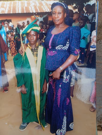 A medium shot captured outdoors shows a young boy and a woman standing side-by-side against a blurred background of people and buildings. The boy, positioned to the left of the frame, wears a green graduation gown and cap with yellow trim. He is looking directly at the camera with a neutral expression. To his right, a woman in a blue and purple patterned dress and headscarf gazes slightly to her left. Her left arm is extended, with her hand resting on the boy's shoulder. The ground beneath them appears to be dirt, with scattered debris. The overall scene suggests a celebratory event, possibly a graduation ceremony, taking place in a community setting in Dutsen Alhaji, Nigeria.