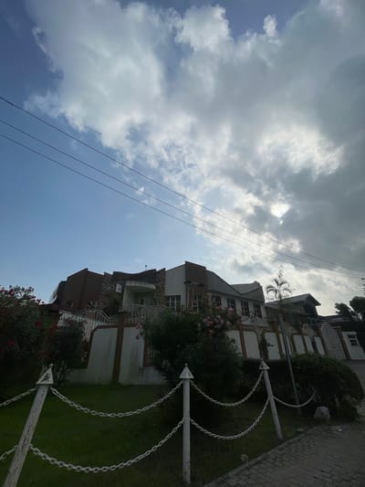 The image depicts a multi-story building in Moba, Nigeria, set against a sky with partly cloudy conditions. Power lines run across the sky. The building appears to have multiple balconies and is bordered by greenery, including trees and flowering shrubs. A white chain-link fence is present in the foreground. The architecture suggests a residential or commercial structure, possibly an apartment complex or office building. The picture is taken from ground level looking up at a slight angle.