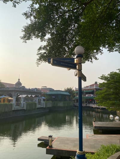 The image depicts a canal-side promenade with buildings and lush greenery. A prominent blue signpost with white lettering, pointing to "LOBBY" and "HACHI GRILL," stands in the foreground, partially obscured by the branches of a large tree. The canal reflects the architecture and the sky, suggesting a serene atmosphere.
Buildings with varied architectural styles line the canal. One building displays the signage "bebek bengil," an Indonesian phrase likely indicating a restaurant. Other structures appear to be commercial establishments or residences, with balconies and outdoor seating areas visible. The presence of umbrellas suggests outdoor dining or a sunny day.
In the distance, people can be seen strolling along the promenade and in the cafes, indicating a leisurely activity or event. The time of day appears to be late afternoon or early evening, with a soft, diffused light and the sky showing a pale hue. The weather seems pleasant, conducive to outdoor activities.
The overall scene conveys a sense of tranquility and a pleasant urban environment, blending natural elements like trees and water with commercial and residential architecture. The reflection on the water adds a dynamic visual element to the static structures. The location, East Jakarta, Indonesia, hints at a tropical climate and a vibrant urban setting.