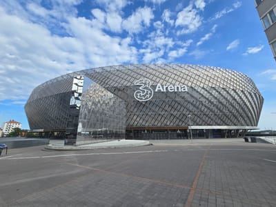 The image displays the Avicii Arena, also known as 3 Arena, situated in the Johanneshov district of Stockholm, Sweden. The dominant feature is the large, spherical arena building, clad in a facade composed of numerous geometric, metallic panels that create a tessellated, reflective surface pattern. The "3 Arena" logo is prominently displayed in white text and symbol on the upper right side of the sphere. Below this, above a series of entry points, the text "14 NOVEMBER AVICII ARENA" and "BENJAMIN INGROSSO" is visible, along with entrance letters 'A', 'B', and 'C'. A significant portion of the lower-left facade consists of large, highly reflective, flat panels that mirror the sky and surrounding structures. The foreground is an expansive, empty paved plaza composed of dark grey rectangular pavers, marked with lighter lines. To the far left in the background, a light-colored residential building with a reddish roof is partially visible. To the far right, a section of a modern, grey building with multiple windows is present. The sky above is clear blue with scattered white cumulus clouds. The scene is brightly illuminated by daylight, and no human figures or discernible activity are present.
