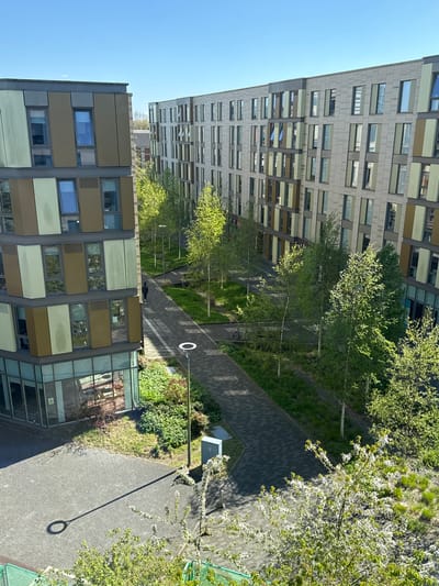An elevated view shows a pathway running between two modern buildings, with landscaping and trees lining the path. The building to the left has a facade of beige and grey panels, while the building on the right is taller with a grey facade. A person is lying on the path between the buildings, with lampposts spaced along the pathway. This image is likely taken in Hull, United Kingdom.