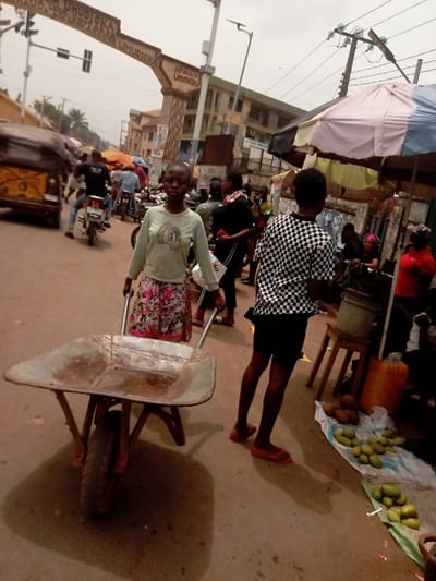 The image depicts a busy street scene in Umudioka, Nigeria, confirmed by a large archway overhead bearing the inscriptions "UMUDIOKA," "APORO CHIEF UMUDIOKA," and "AKWA ADAGHA ANAMBRA STATE UMUDIOKA." The sky is overcast.
In the foreground, a young individual with short hair, wearing a light green long-sleeved shirt with a dark "B" graphic and a floral skirt, directly faces the viewer while pushing an empty, rusty metal wheelbarrow with a single front wheel. To the right, a second individual is seen from behind, wearing a black and white checkered short-sleeved shirt, black shorts, and red flip-flops, standing near a market stall.
The market stall, partially visible on the right, operates under a multi-colored umbrella (pink, light blue, yellow segments). On the ground, green mangoes or similar fruit are displayed on a white plastic sheet, alongside several brown coconuts. A wooden stool supports a grey bucket, and a large orange plastic container rests nearby.
The background shows a bustling street with multiple pedestrians and vehicles. A yellow motorized tricycle is partially visible on the far left. Several motorcycles, some ridden by individuals, are present in the mid-ground. Multi-story buildings line the street, with one displaying a sign partially legible as "FELIX ZINGA BANK." Overhead, utility poles with power lines and traffic signals are visible. The street surface appears to be rough, unpaved, or heavily worn asphalt.