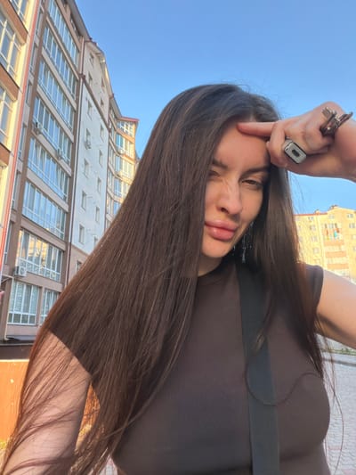 The image is a selfie taken outdoors, possibly in an urban environment. The subject is a woman with long, dark brown hair, wearing a brown, ribbed, sleeveless top. She is touching her forehead with her right index finger, and her right hand is holding several keys and a ring with a dark object. Her lips are slightly pursed, and she is looking towards the camera with her left eye partially closed. In the background, there are buildings with multiple stories and windows, suggesting an apartment complex or similar structure. The sky is clear and blue. Based on the location information, this scene is likely in Vovchynets, Ukraine.