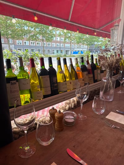 This is a photograph of a table set up in what appears to be an outdoor seating area of a restaurant. A red awning is visible on the left side of the image, suggesting it might be a covered patio or balcony. Through a window or glass partition behind the table, a city scene is visible, featuring buildings with many windows and trees with green foliage, indicative of daytime. The buildings and trees suggest an urban environment, possibly Copenhagen, Denmark, given the context provided.

The table is laden with several wine bottles, mostly red and white, arranged in a rack or on a slanted surface. Various wine glasses are also present, some empty, one partially filled, and others neatly placed. A small candle in a glass holder and a salt and pepper grinder are also on the table, suggesting a dining setting. Scattered corks add to the ambiance of a wine-focused establishment. A fork and a napkin are also visible, hinting at a meal in progress or about to begin. The overall mood seems relaxed and inviting, hinting at a pleasant dining experience.

Visible text on the wine bottles includes brand names and descriptions such as "RIOJA Baroja CRIANZA 1988," "Cuve Sa 2020," "7th Class Cabernet Sauvign," "MOUTON-CAL Baron Philippe," "DOMAINE DES CHA," "MEURSAULT COTE D R," "RULLY," and "INTERNATIONAL HOUSE." The lighting from decorative bulbs strung across the awning suggests the photograph might have been taken in the late afternoon or early evening.