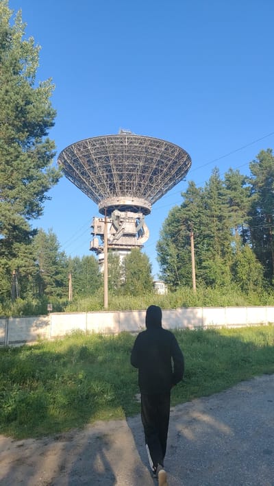 The image depicts a large radio telescope against a clear blue sky, located in the vicinity of Tolstoukhovo, Russia. In the foreground, a person wearing a black hooded jacket and black sweatpants with white stripes is walking away from the camera on a dirt and asphalt path. The person casts a shadow on the ground. To the left and right of the radio telescope are tall green pine trees. A concrete wall runs horizontally across the middle ground, separating the path from an area of overgrown green grass and bushes. The radio telescope itself is a massive, intricate metal structure supported by a tall concrete and metal tower. The overall environment appears to be rural or industrial, with elements of nature interspersed with large technological infrastructure.