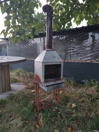 The image depicts an outdoor scene, likely in a backyard or garden, in Kochmar, Bulgaria. The central object is a metal barbecue grill with a tall chimney. The grill appears to be old and somewhat rusty, with a weathered metal construction. It is supported by a red metal frame. To the left of the grill, there are lush green leaves of a tree, obscuring part of the background. A brown pipe, possibly part of the chimney system or a gutter, is visible in the upper left corner.
Behind the grill, there is a structure covered with a translucent, crinkled material, possibly a plastic tarp or sheeting, giving it a somewhat abstract appearance. The ground around the grill is covered in grass and dry leaves, suggesting it might be autumn. There are also some wooden elements in the foreground, including what looks like a section of a large wooden cable spool and some wooden planks.
The lighting suggests it is daytime, with a soft, diffused light, perhaps indicating an overcast sky or late afternoon. There are no people visible in the image, and the overall impression is one of a quiet, slightly neglected outdoor space. The scene is static, with no specific activity taking place. The location cues, such as the mention of Kochmar, Bulgaria, and the rustic setting with outdoor cooking equipment, provide context.