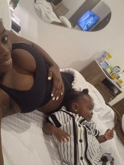 An overhead-angle image, rotated counter-clockwise, shows an adult and a young child lying on a bed. The adult, who appears to be a dark-skinned woman, is positioned in the upper right of the frame (as oriented in the original image). She is wearing a form-fitting black top or sports bra that accentuates her cleavage. Her face is visible, showing a slight smile. Her left hand, with white nail polish, rests on her lower abdomen.

To her left, a young child, also dark-skinned and possibly a toddler, lies on their back. The child is wearing a white long-sleeved shirt with black horizontal stripes and small dark buttons down the front. A small gold earring is visible in the child's right ear. Their head is turned slightly away from the camera, and their expression is neutral. Both individuals are lying on white bedding.

In the background, towards the bottom right of the frame (as oriented in the original image), a large, oval-shaped mirror reflects a portion of the room. Within the reflection, a screen, possibly a television or tablet, displays a brightly lit scene with a blue hue, resembling an animated show or perhaps an aquarium. Next to the mirror, a wooden nightstand or small cabinet is visible, holding various small bottles and containers, suggesting toiletries or personal items. No discernible text is visible in the image. The scene conveys a relaxed, intimate moment between the adult and child.
