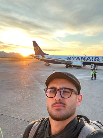 A man is taking a selfie in front of a Ryanair airplane on the tarmac. The photo was likely taken at Orio al Serio Airport in Italy, as Ryanair is a common airline there. The sun is setting or rising in the background, casting a warm glow on the scene. The man is wearing glasses and a hat. He is dressed casually with a backpack. In the background, there is an airplane, a Ryanair plane, airport staff, and a ramp with safety cones. The ground is paved.