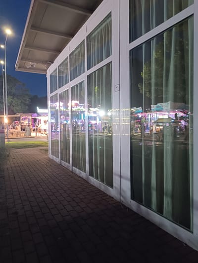 The image depicts a nighttime scene in Viadana, Italy, likely a fairground or amusement park area. In the foreground, a paved walkway made of dark interlocking bricks leads towards a modern building with large glass windows and white frames. The windows are covered by sheer, light-colored curtains, which reflect the brightly lit amusement park attractions visible in the background. These attractions appear to be rides, possibly a carousel or similar structures, illuminated with colorful lights. There are a few indistinct figures visible through the glass and in the distance within the park. The sky above is dark blue, indicating it is nighttime, and streetlights are visible on the left side of the image. The overall impression is one of a vibrant, illuminated entertainment area after dusk.