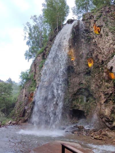 The image shows a waterfall cascading down a rocky cliff face. The cliff is covered in moss and vegetation, and there are several glowing orange lights embedded in the rock, illuminating some of the crevices. Trees grow at the top of the cliff and on the surrounding slopes. At the base of the waterfall, there is a pool of water and a rocky shoreline. In the foreground, a wooden bench is partially visible. The sky is overcast. The location is identified as a waterfall in Krasny Kurgan, Russia. There are no people visible in the image.