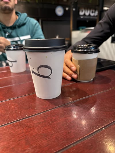 Two coffee cups sit on a red-brown wooden surface. The first cup has a large black "9" on it, with a black lid. The second cup has a similar design but the number is obscured. A hand holds a third cup with a brown sleeve. Part of a person's face is visible in the bottom left, wearing a green and white top. Another person's arm, clad in black, is partially seen at the top left. The background appears to be a coffee shop interior.
