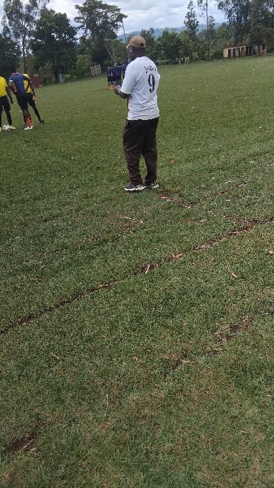 The image depicts a man in a white shirt with the number "9" on the back standing on a grassy field. He is facing away from the camera and appears to be holding a remote control in his hand.  A small group of people are visible in the background, standing in the distance on the same grassy field. The scene is a soccer field in Bungoma, Kenya, located at latitude 0.554, longitude 34.774. The field is covered in green grass with some patches of dirt visible. There are several visible tire tracks on the field. The image is taken from a low angle, making the man appear tall in the frame. The weather appears to be overcast, with a dull, grey sky in the background.