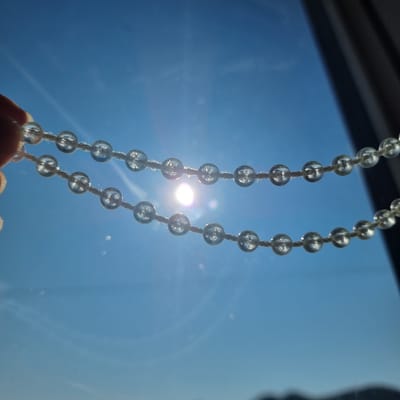 The image shows two strands of clear, spherical beads against a bright blue sky with a sunburst. A hand is visible on the left, holding the upper strand of beads. The beads are strung on a white cord. The sun is positioned centrally between the two strands, creating lens flares and highlights on the beads. In the upper right portion of the image, a dark, vertical object, possibly a window frame or curtain, obscures part of the sky. The lower part of the image shows a blurred, dark silhouette of a mountainous landscape. The overall scene suggests an outdoor view from a window on a sunny day.