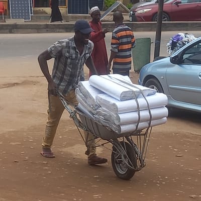 The image depicts a street scene in Sardauna, Nigeria. A man in a blue baseball cap, plaid shirt, and khaki pants is pulling a wheelbarrow loaded with tightly packed, white rolls, possibly paper or fabric, secured with ropes. Two other men are nearby. One is wearing a red robe and a white cap, and the other is wearing a horizontal striped shirt. Various objects are in the background, including cars, a green patch of grass, and what appears to be a display of solar panels. The ground is a light brown color, likely dirt or pavement.
