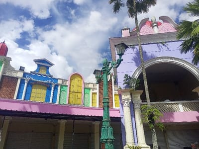 The image depicts a street scene in Gadog, Indonesia, characterized by colorful, European-inspired architecture under a bright blue sky with white clouds.
**Subjects and Objects:**
The foreground features an ornate, dark green lamppost with intricate scrollwork and a traditional lantern head, positioned slightly left of center. To its right, two light-colored, fluted classical columns with Corinthian capitals support an upper structure. A slender palm tree trunk leans diagonally across the right side of the image, with green fronds visible.
The scene is dominated by a row of multi-story buildings, appearing to be commercial or retail structures. The building on the left exhibits a mix of architectural elements, including a reddish-brown textured facade at the lower level, topped by a pink roofline. Above this, a blue section features a central pediment-like element over a yellow panel, flanked by green and white decorative details, and an arched yellow panel to its right.
The building on the right presents a large purple facade with a prominent white arched opening on its upper level, above which is a decorative pink gable roof with white scrollwork and a finial. A white lattice railing is visible below the arched opening. Below the pink roof overhang, the ground level of both buildings is uniformly closed with multiple light gray, roll-down security shutters, indicating inactive storefronts or a closed establishment.
**Environment:**
The sky is clear blue with scattered white cumulus clouds, suggesting daylight conditions. Strong natural light creates defined shadows on the buildings. No individuals, vehicles, or overt activities are visible, presenting a static and unoccupied scene.