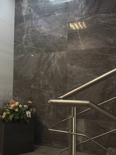 The image shows a close-up of a dark gray marble tiled wall with a modern stainless steel railing. The railing consists of several curved pipes and supports, reflecting the ambient light. In the lower right corner, a dark rectangular object, possibly a planter or a box, holds a bouquet of artificial flowers in various colors, including white, pink, and yellow, with green foliage and delicate branches. The lighting creates a somewhat dramatic effect with highlights on the polished surfaces of the railing and the marble. Some areas of the marble show natural veining. A reflection of what appears to be light fixtures is visible on the left side of the wall. The scene suggests an indoor setting, likely a staircase or a hallway, with a contemporary and somewhat luxurious aesthetic. There is no visible text. The time of day is not discernible, and the weather is irrelevant as it's an indoor scene. The location is given as Mahilyow, Belarus.