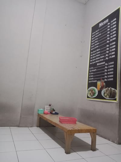 The image captures a simple dining area with a wooden bench and a menu board displayed on the wall. The bench, made of unfinished wood, is positioned in the lower half of the frame, with a pink plastic basket resting on its surface. A white tiled floor extends across the foreground.
On the right side of the image, a black menu board is attached to a light gray wall. The menu is written in white text and lists various food and drink items with their prices. Below the food items, there is a section for "Minuman" (Drinks). The menu board also features a small graphic at the bottom displaying two bowls of food and two glasses of drinks.
On the bench, to the left of the pink basket, are several small containers, including a white patterned holder with what appear to be utensils or chopsticks, a light blue rectangular box, and a pink cup. A small blue tray is also visible on the bench.
The walls are painted a neutral gray, with a vertical seam or joint visible on the left side of the frame. The lighting appears to be artificial, casting shadows on the floor. The overall impression is of a modest, functional eating space.
The user indicates this location is in the city of Cangkring, Indonesia. This suggests a small, local establishment, likely a warung or small restaurant, common in Indonesian towns and cities. The simplicity of the furniture and the style of the menu are consistent with this type of establishment.
