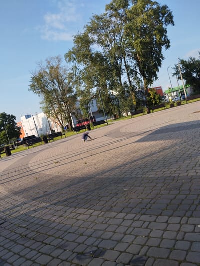 The image captures an outdoor scene on a bright, sunny day with a clear blue sky and scattered clouds. The foreground is dominated by a paved plaza made of interlocking gray bricks, arranged in concentric circular patterns. Sparse green grass and dry leaves are visible between the bricks. In the middle ground, a person wearing a dark blue hoodie and light-colored pants is crouched down on the pavement, seemingly interacting with something on the ground. To the left and behind the person are several dark-colored benches and trash receptacles. Further in the background, a line of trees with green foliage stands tall, casting shadows across the plaza. Beyond the trees, buildings are visible, including a white building with dark windows and some industrial-looking structures with colorful signage, one featuring the Cyrillic word "СТРОЙМАТЕРИАЛЫ" (STROIMATERIALY), which translates to building materials, suggesting a commercial area. Streetlights are also present. The overall environment appears to be a public space, possibly a park or a square adjacent to a commercial zone in the city of Kingisepp, Russia.