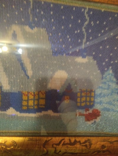 The image is a close-up, slightly blurry, and overexposed photograph of a framed artwork, possibly a diamond painting or cross-stitch. The artwork depicts a winter scene with a dark blue, starry sky. Below the sky, there are several elements that suggest a holiday or festive theme. On the left side, there appears to be a white structure with a blue accent, possibly a house or a snowman. In the center and to the right, there are two gift-wrapped packages. The one on the left is yellow with red ribbons, and the one on the right is also yellow with red accents. To the far right, there is a stylized blue Christmas tree. In the foreground of the artwork, there is a patch of light blue with some darker blue and white areas, suggesting snow or a building. At the very bottom of the artwork, there is a red, heart-shaped object with white accents, which could be a decoration or a stylized figure. The artwork is housed in a golden frame with a textured surface. The lighting in the photograph is harsh, causing glare and obscuring some details of the artwork. There are no identifiable landmarks or features that place this image specifically in Chernihiv, Ukraine.