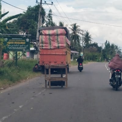 This image depicts a rural or semi-rural roadside scene in Gampongrayadagang, Aceh, Indonesia, under an overcast sky during daytime.

In the foreground, a large, red truck is parked or stopped on the left side of an asphalt road. Its cargo is covered with a striped, multi-colored tarp (red, green, beige). A small, wooden, four-legged stand is positioned directly in front of the truck's rear. To the left of the truck, a person is seated on the grassy roadside verge.

A green sign stands prominently on the left side of the road, partially surrounded by banana leaves and other foliage. The sign clearly displays "RUDI MUSLIMAT" in Latin script, and below it, "المدرسة المعهد الإسلامية ردي مسلمات" in Arabic script, which translates to "Islamic Institute Rudi Muslimat." Further text on the sign indicates an address: "B.B. Anom - Munder Km. 20.7 Desa Pante Pirak, Kec. P. R. gangan Kab. Bireun Prov. Aceh." A red gas tank is visible behind the sign.

The road itself is asphalt and curves gently to the right. Two motorcyclists are seen driving away from the camera. The motorcyclist in the foreground is wearing a maroon or brown head covering (hijab), while the one further down the road is dressed in black. The background is filled with lush green vegetation, including palm trees, indicative of a tropical environment. Utility poles and power lines crisscross the upper part of the image against the cloudy sky.